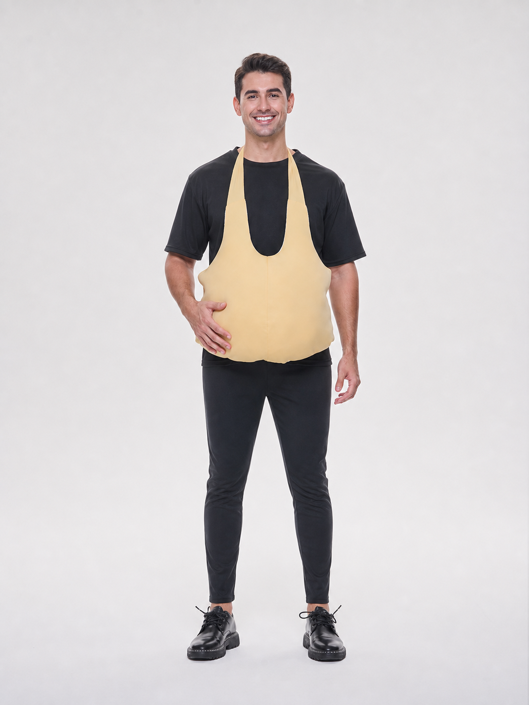 Wearable fake belly costume accessory with a large rounded shape. The model has a bib-style front panel, two shoulder straps, and side ties for fastening. The smooth solid-color surface comes in a light beige or beige shade, so the accessor