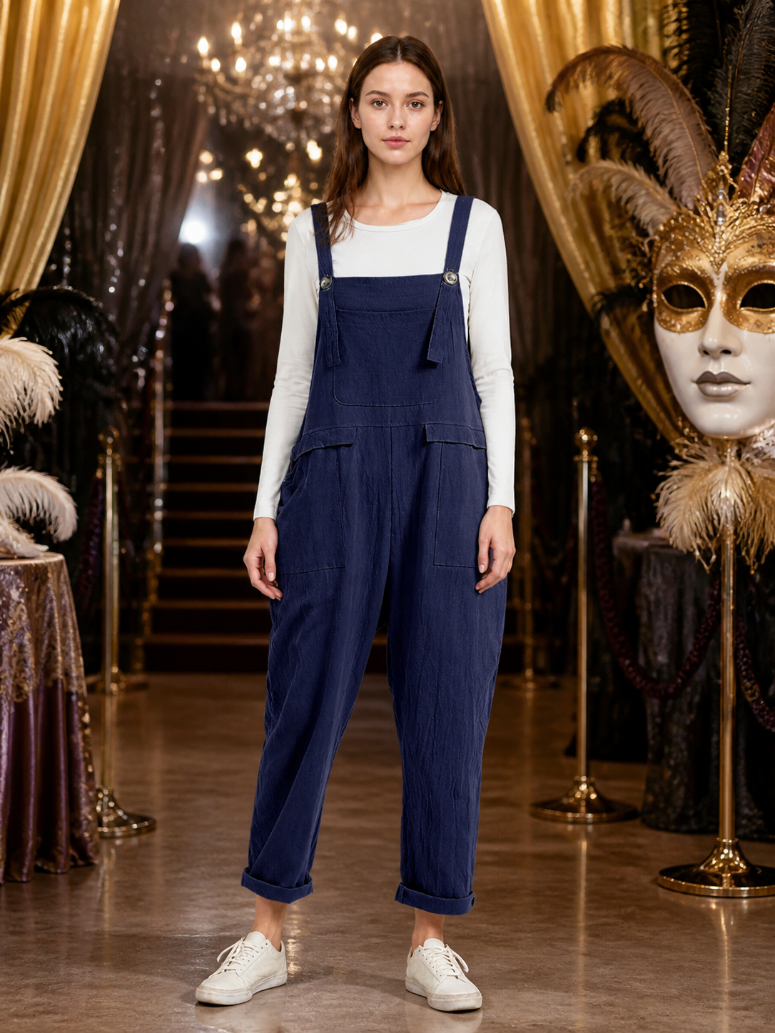 Front view of navy blue bib overalls over a white long-sleeve shirt, styled with white sneakers in an ornate indoor setting