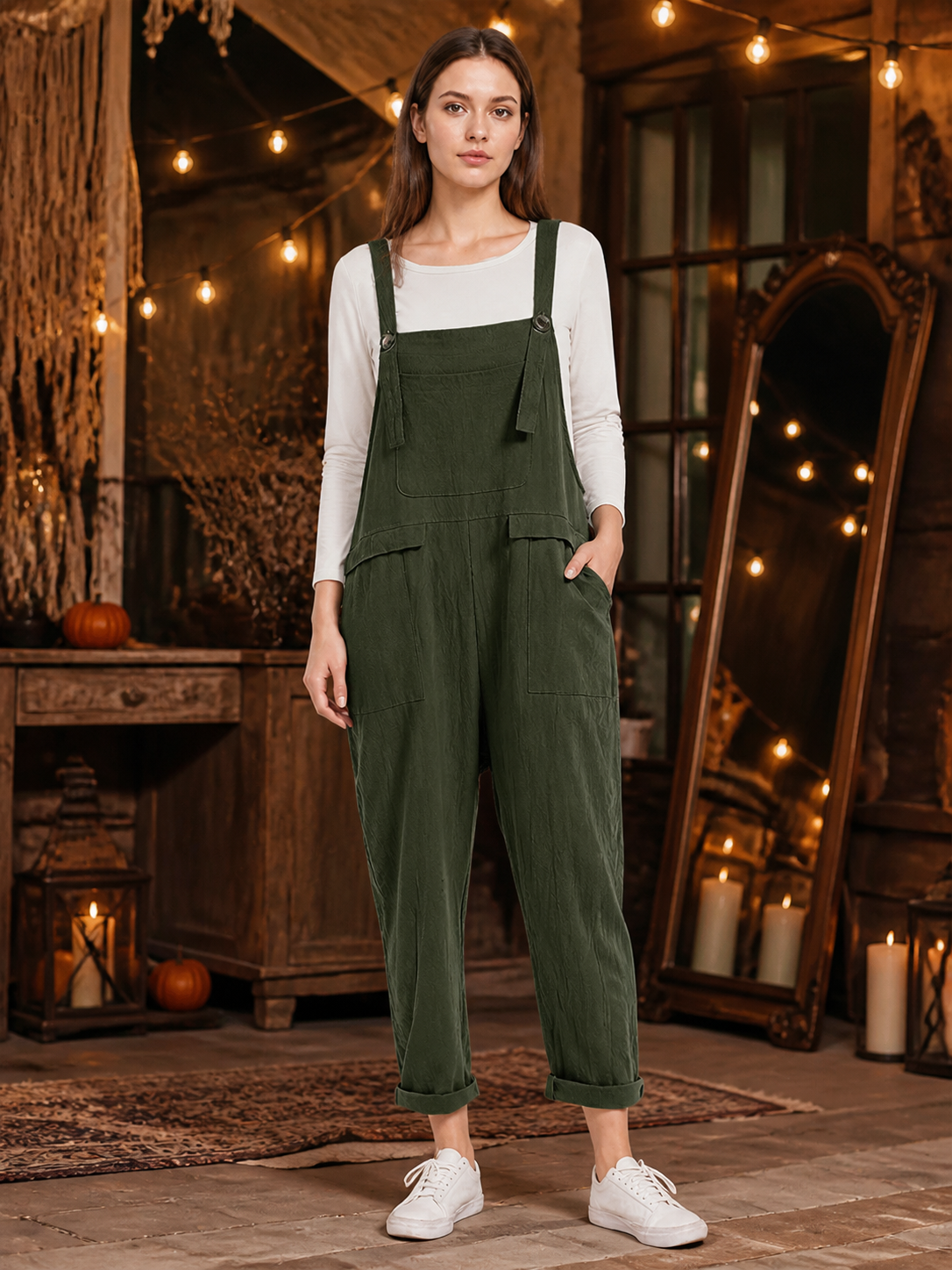 Full-body model wearing dark green textured bib overalls with a chest pocket, side pockets, rolled cuffs, and white sneakers in a rustic room