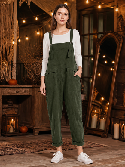 Full-body model wearing dark green textured bib overalls with a chest pocket, side pockets, rolled cuffs, and white sneakers in a rustic room