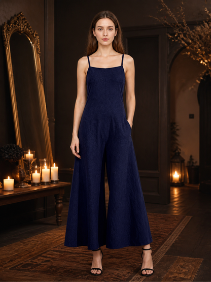 Navy blue wide-leg jumpsuit with thin straps and side pockets shown full-length on a model in a dim room