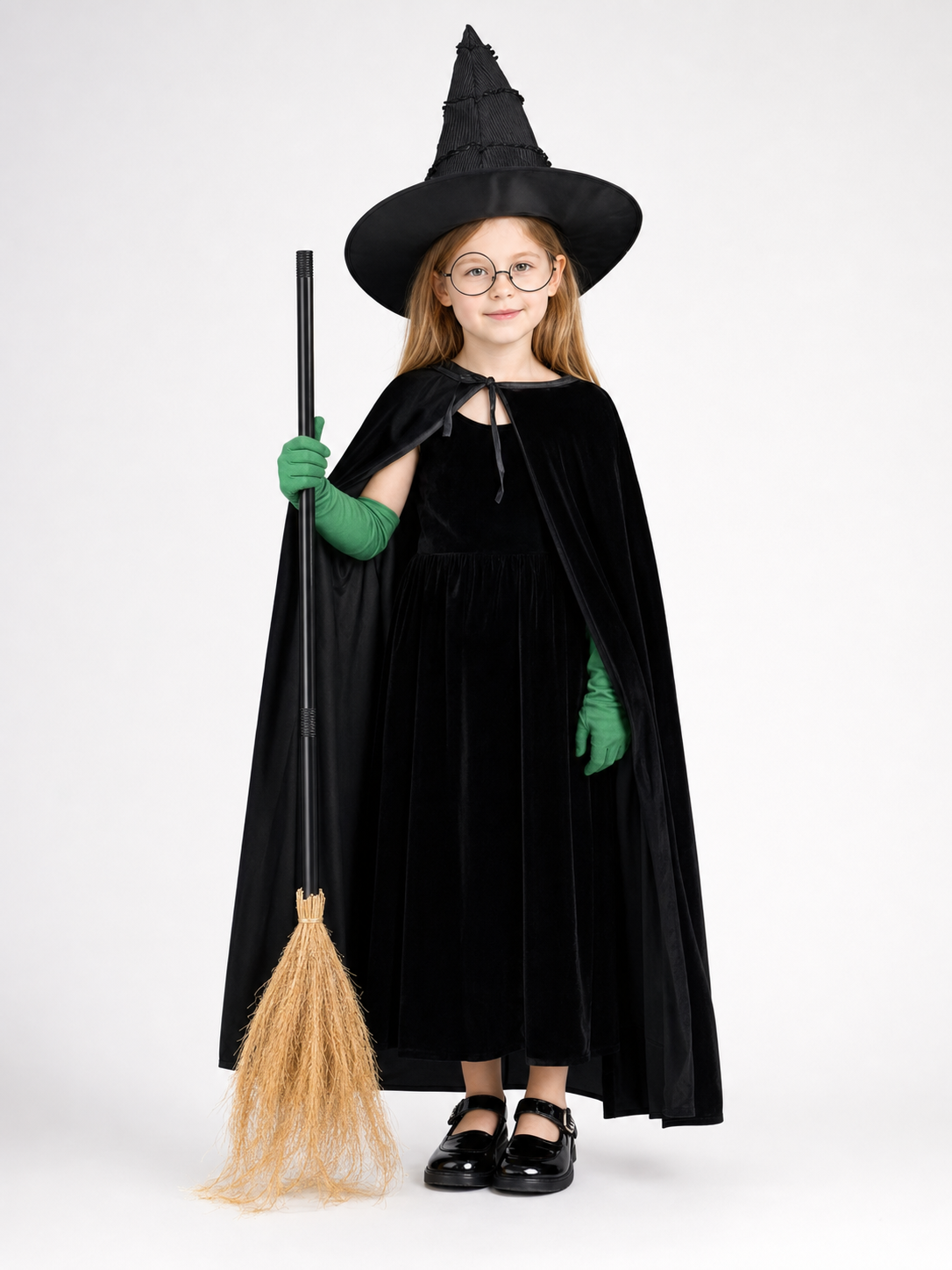 Full-body child model wearing a black velvet dress and cape, pointed hat, round glasses, green gloves, and holding a broom 2