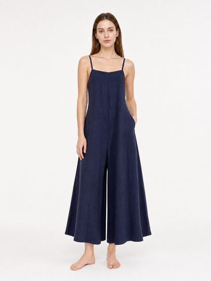 Dark blue wide-leg jumpsuit with thin straps and side pockets, shown full-length on a barefoot model against a light background