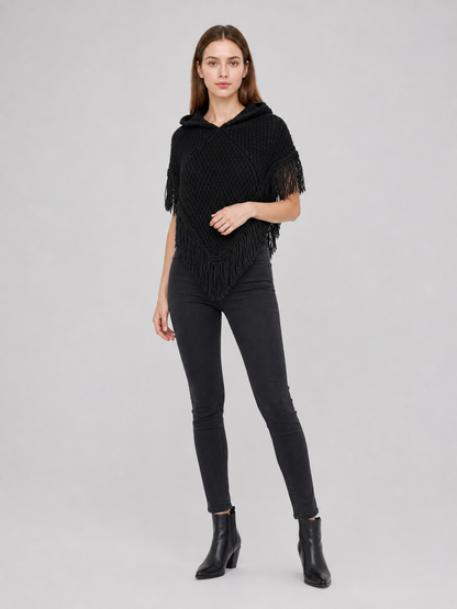 Black hooded knit poncho with pointed fringe, shown on a model wearing jeans and ankle boots against a gray background