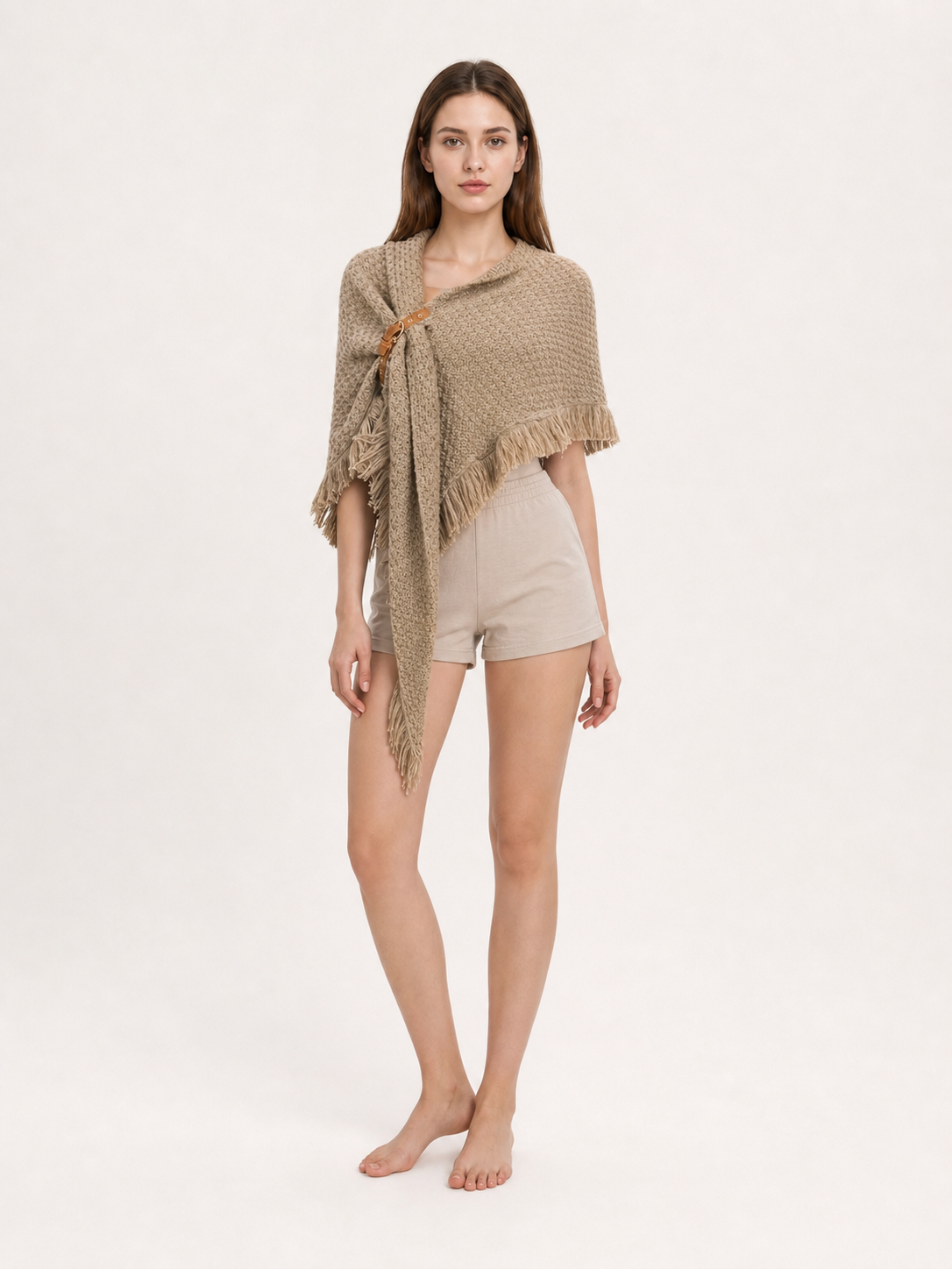 Beige fringed cape styled with light shorts, shown full-body against an abstract cream background
