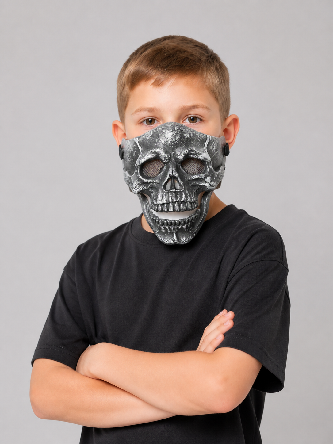 Black skull face mask with hollow eye openings and layered molded teeth, shown front-facing on a child with folded arms