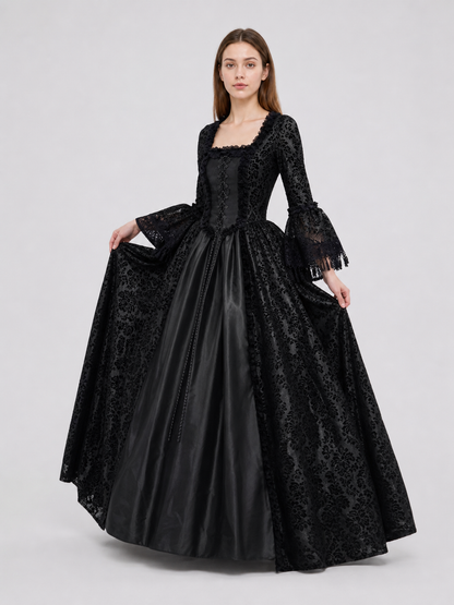 The black dress is designed in the aesthetic of a Victorian-era Gothic costume. The model features a floor-length silhouette, a fitted bodice with a square neckline, and decorative lacing or trim on the front. The long flared sleeves end in