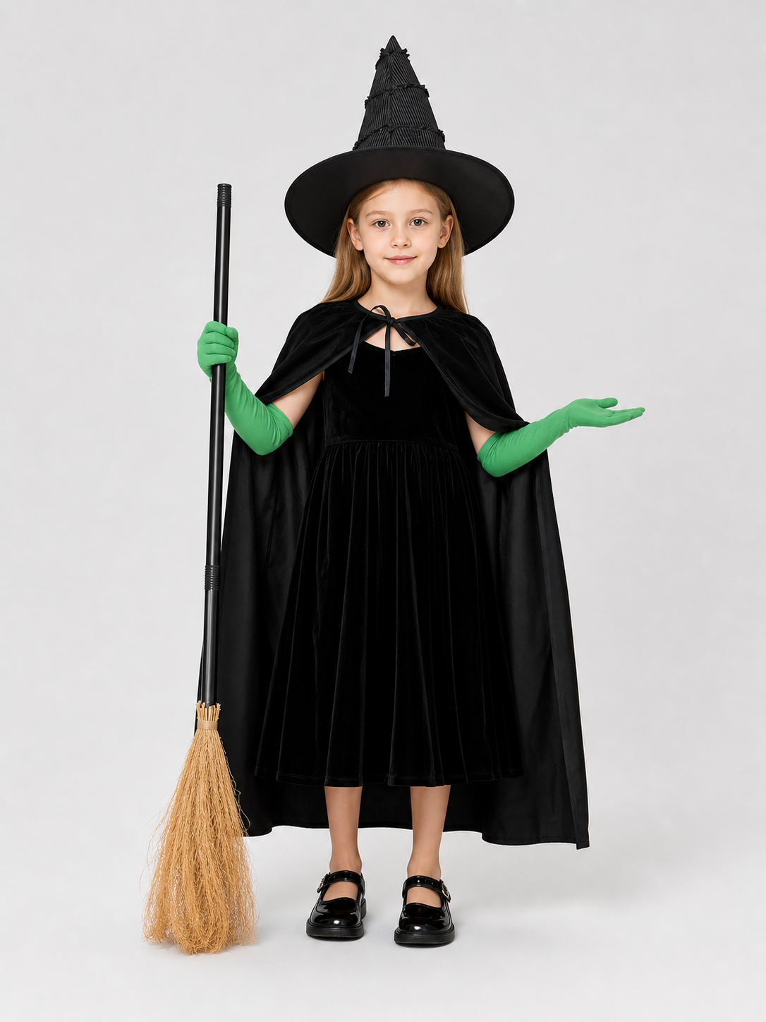 Full-body child model wearing a black dress, cape, pointed hat, and green gloves, holding a broom with one hand raised