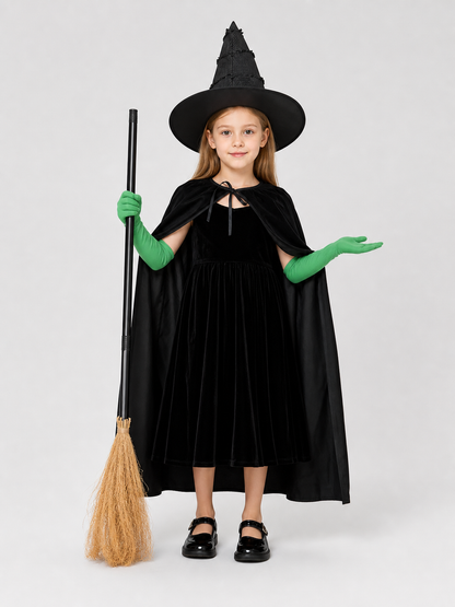Full-body child model wearing a black dress, cape, pointed hat, and green gloves, holding a broom with one hand raised