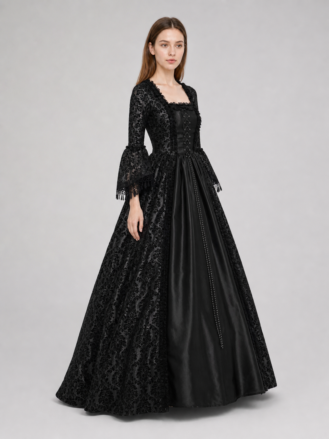 The black dress is designed in the aesthetic of a Victorian-era Gothic costume. The model features a floor-length silhouette, a fitted bodice with a square neckline, and decorative lacing or trim on the front. The long flared sleeves end in