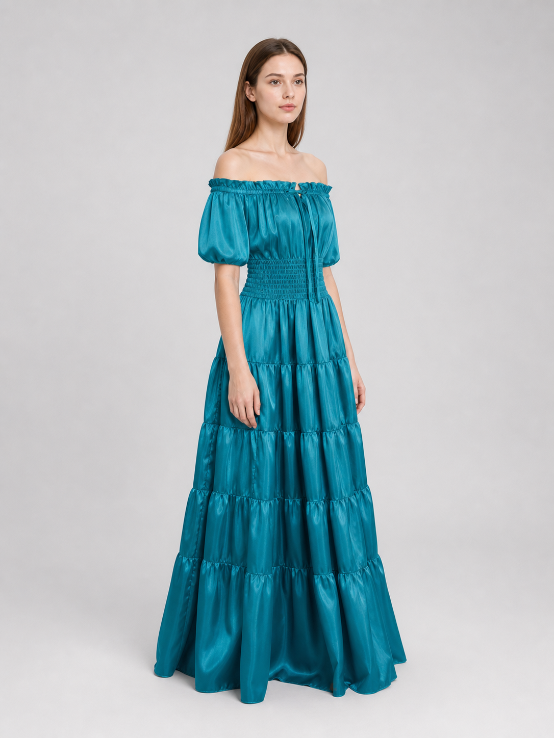 Three-quarter side view of a teal maxi dress on a model, showing its glossy fabric, short puff sleeve, smocked waist, and long tiered skirt