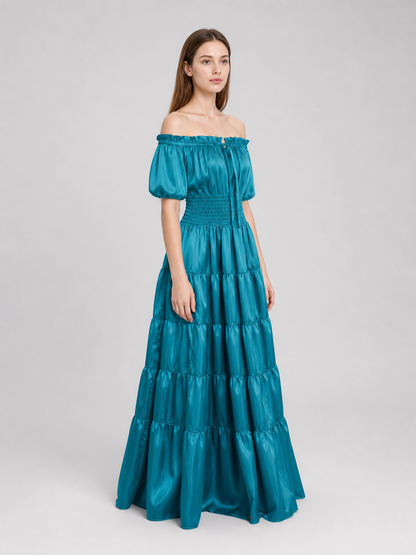 Three-quarter side view of a teal maxi dress on a model, showing its glossy fabric, short puff sleeve, smocked waist, and long tiered skirt