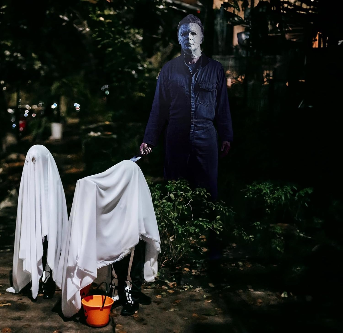 Life-size Michael Myers horror cutout standee in a dark yard behind two kids dressed as ghosts