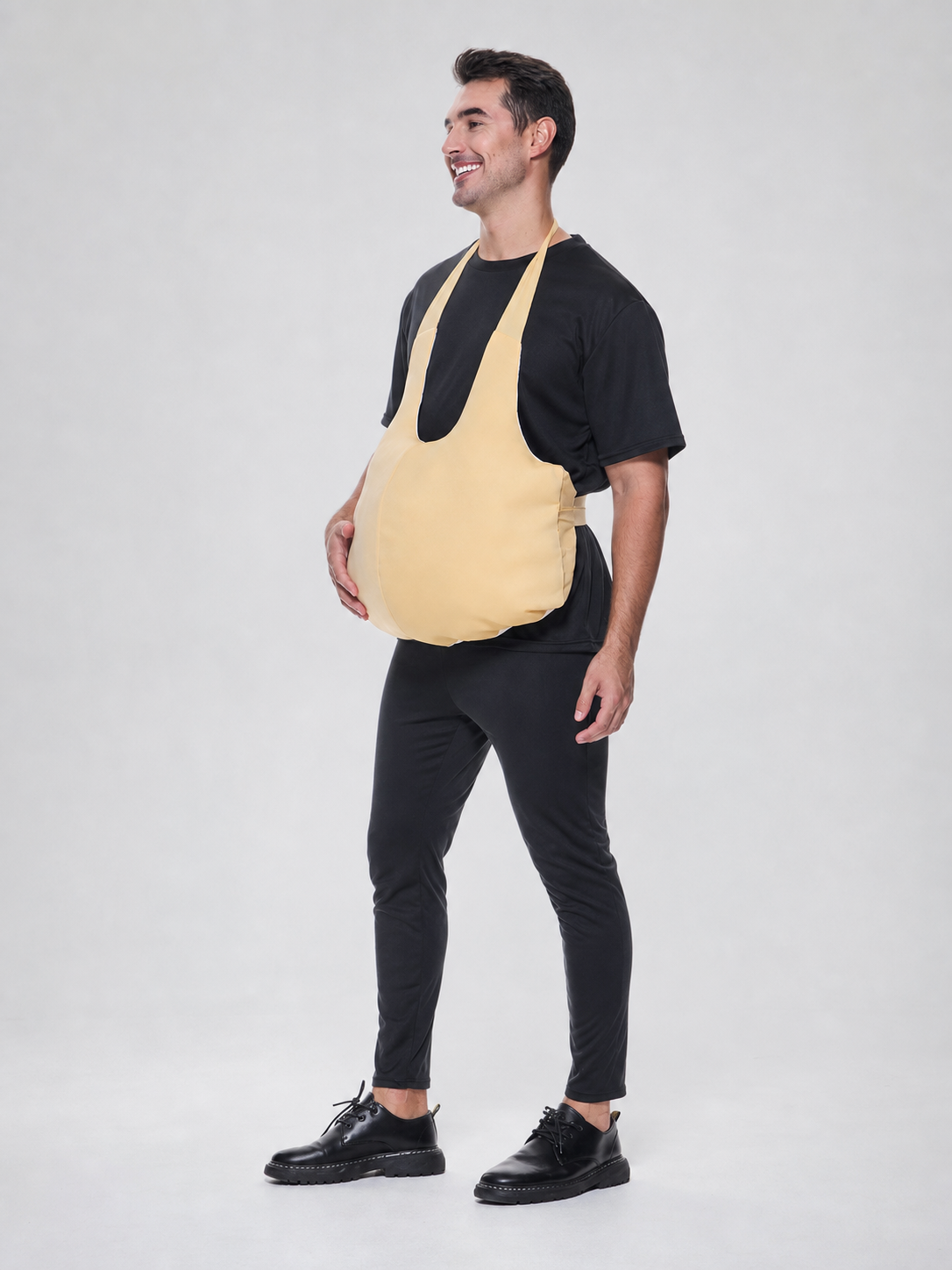 Wearable fake belly costume accessory with a large rounded shape. The model has a bib-style front panel, two shoulder straps, and side ties for fastening. The smooth solid-color surface comes in a light beige or beige shade, so the accessor