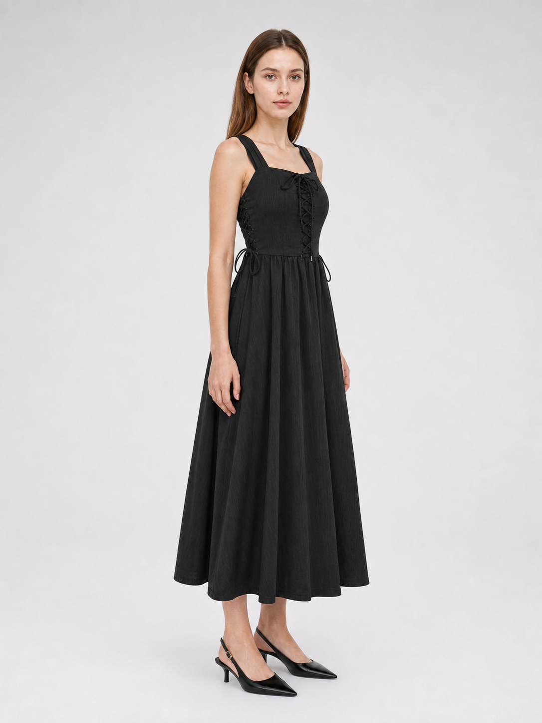 Side view of a black midi dress on a model, showing the fitted bodice, wide shoulder straps, side waist tie, and long flared skirt
