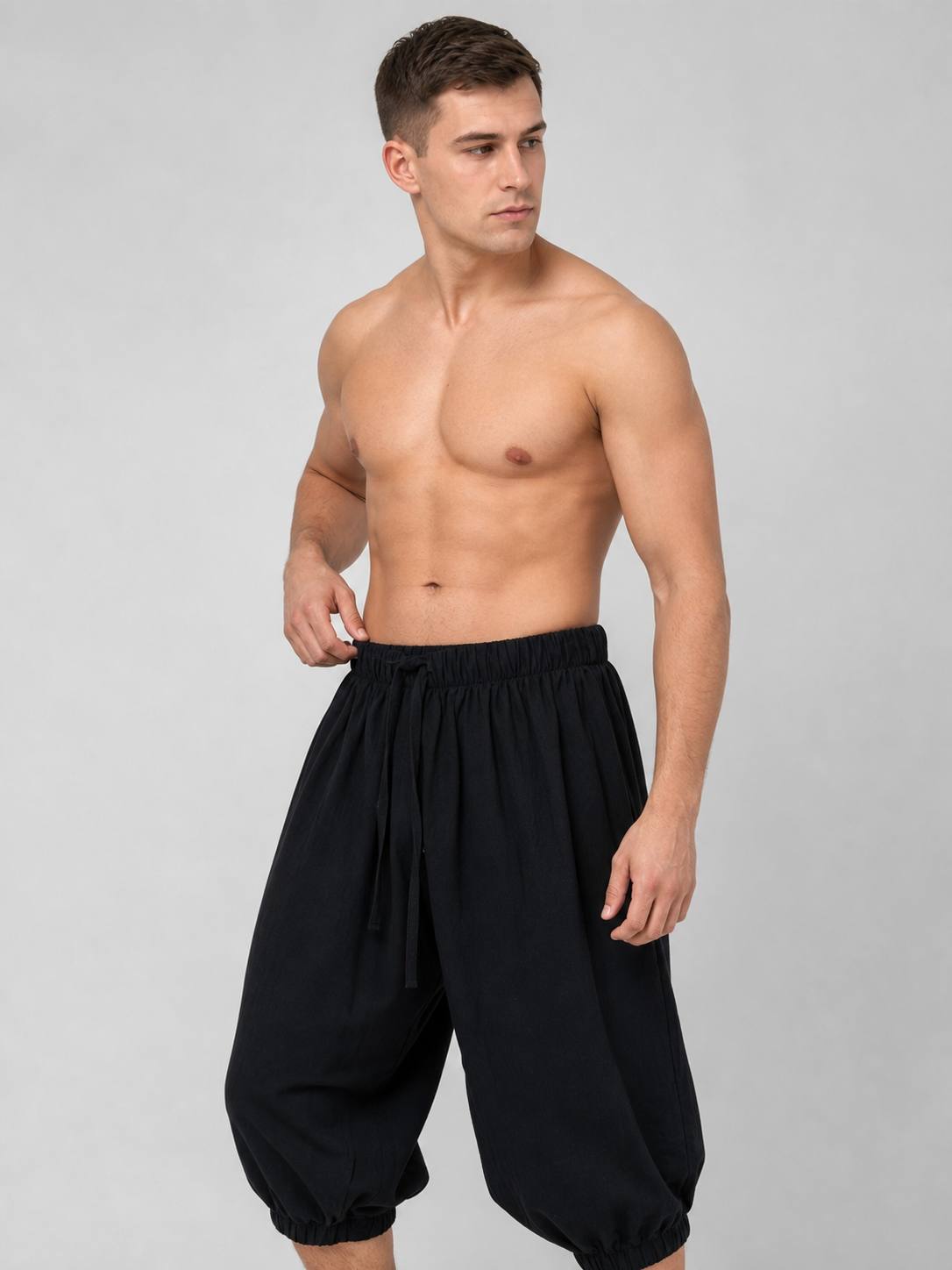Angled view of black cropped pants on a shirtless model, showing the elastic waist, drawstring, and gathered ankle cuffs