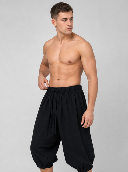 Angled view of black cropped pants on a shirtless model, showing the elastic waist, drawstring, and gathered ankle cuffs